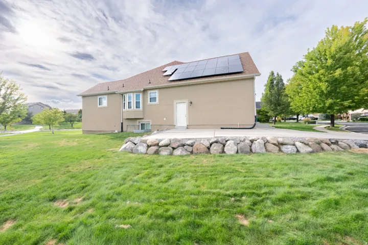 Exterior featuring solar panels, a neutral-toned stucco finish, multiple windows, a rear patio, and a stone retaining wall