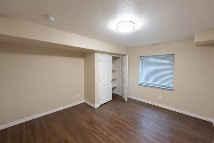 Wood-finish flooring, recessed window with blinds, built-in closet with shelving, overhead flush-mount lighting, and neutral wall paint
