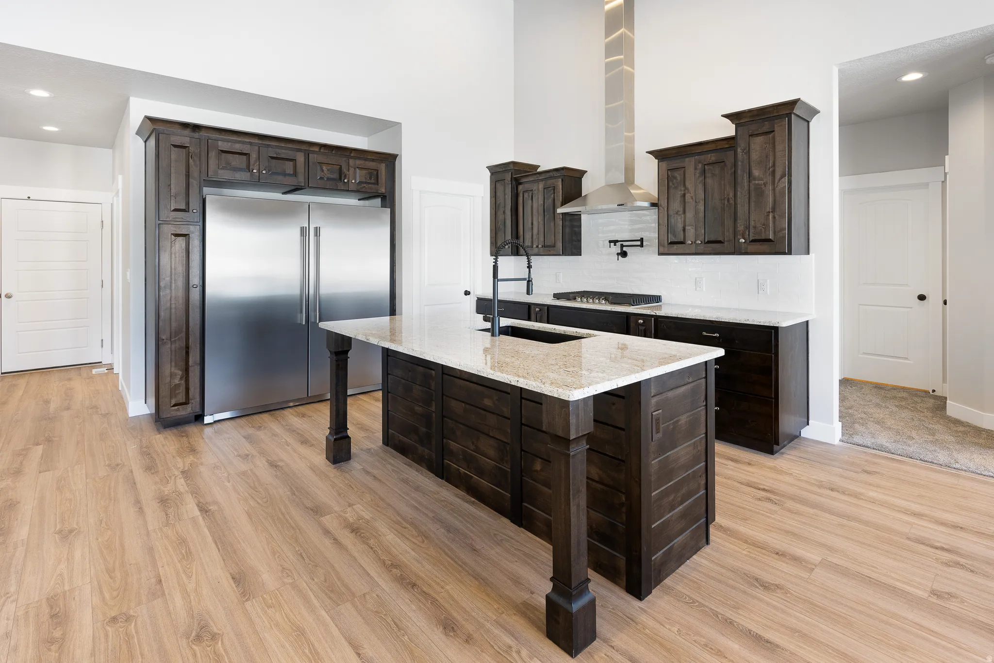 Kitchen featuring an expansive island with a light-toned countertop, stainless steel appliances, dark wood-finish cabinetry, a white tile backsplash, and wood-finish flooring