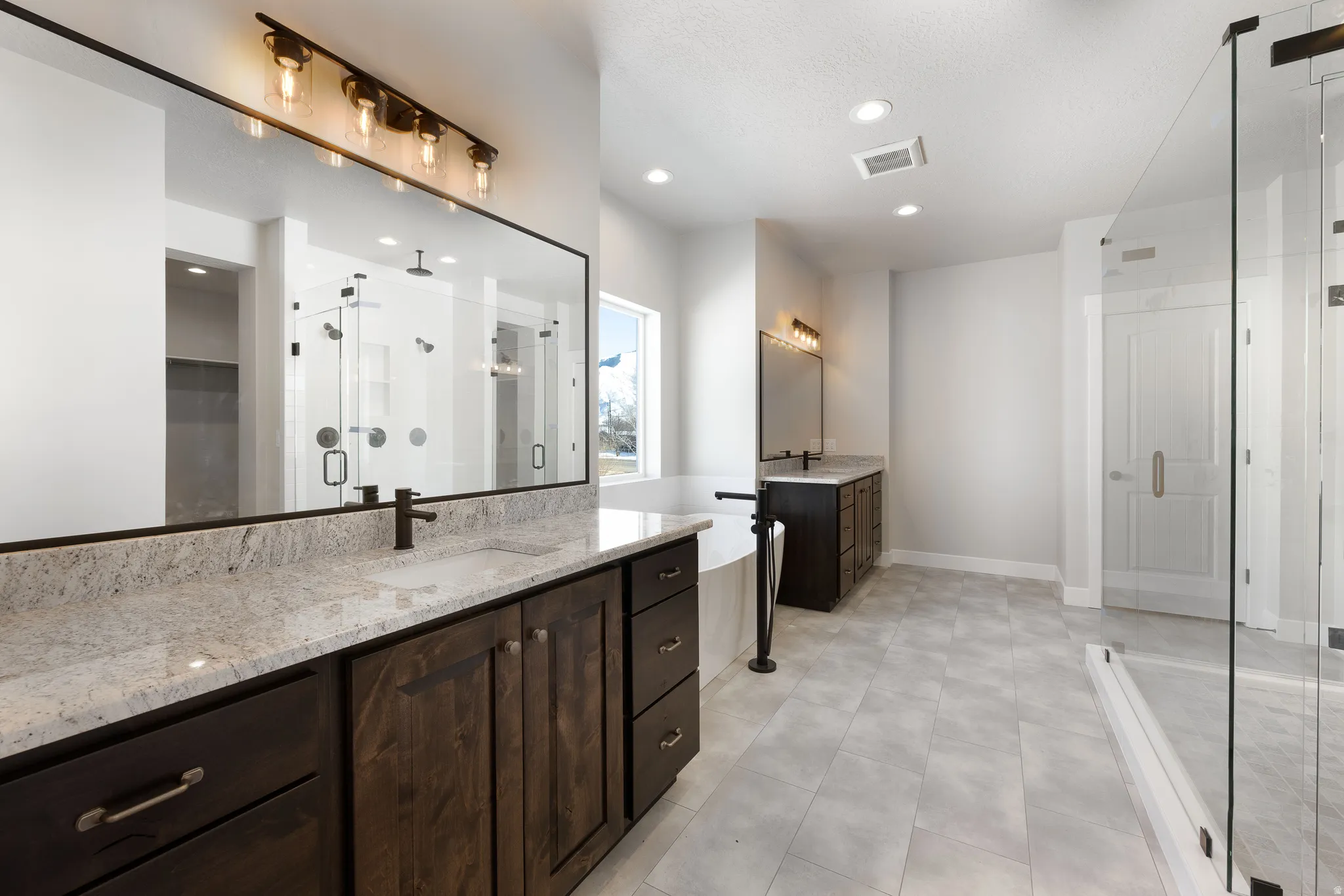 Dual dark wood-finish vanities with light speckled countertops, a freestanding tub with a floor-mounted faucet, a spacious walk-in shower with a glass enclosure, and light gray tile flooring