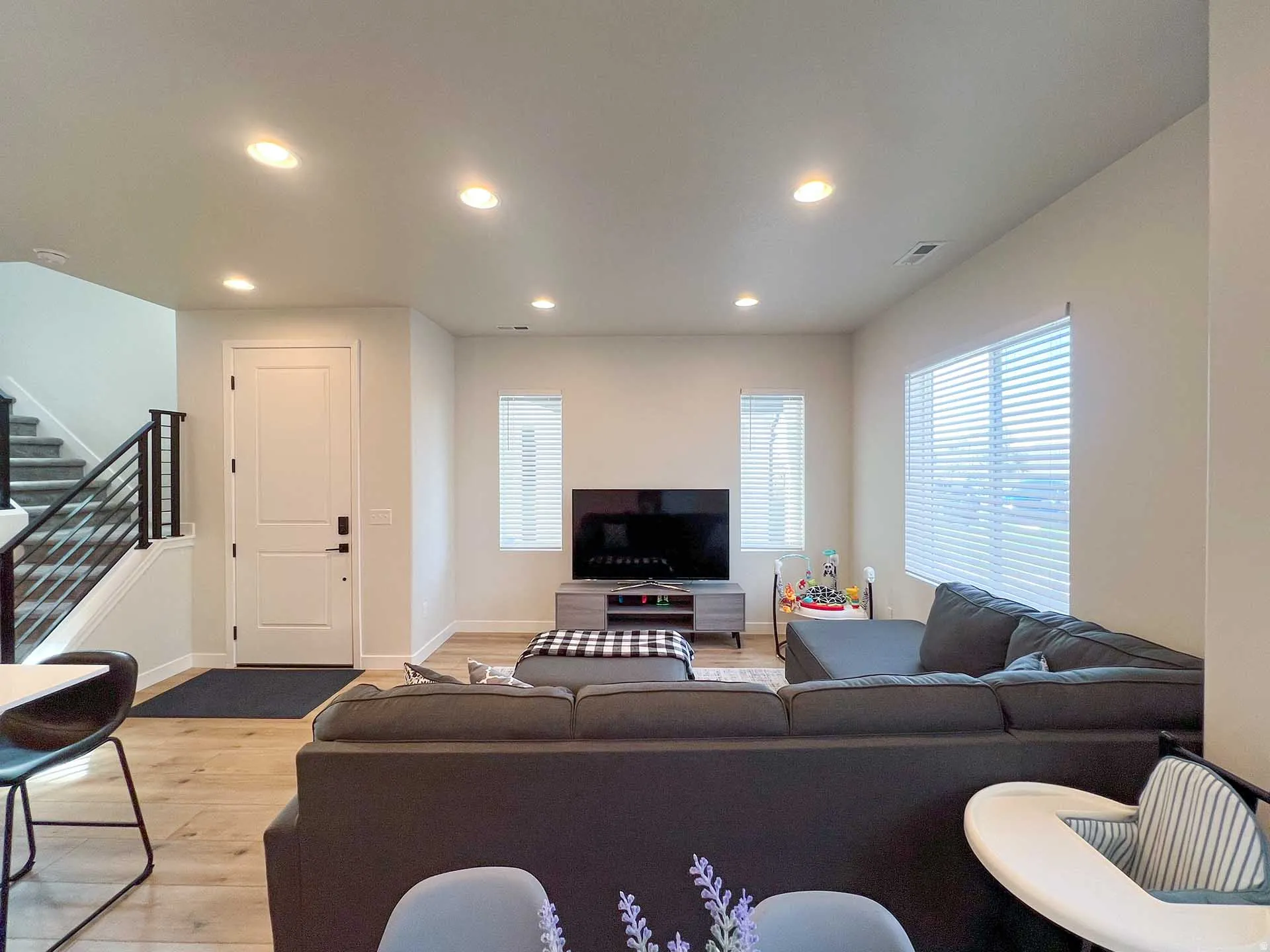 Recessed ceiling lighting, light wood-finish flooring, staircase with black metal balusters, white interior door, and multiple windows with blinds