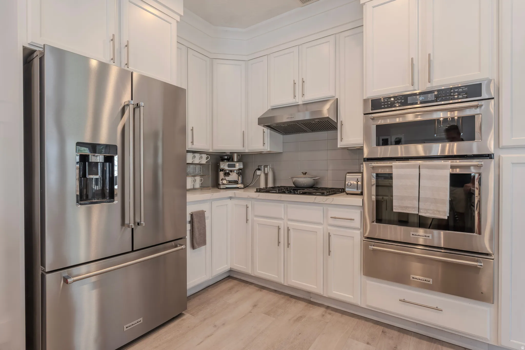 White shaker-style cabinetry, stainless steel appliances, light-toned wood-finish flooring, white countertops, and a tiled backsplash