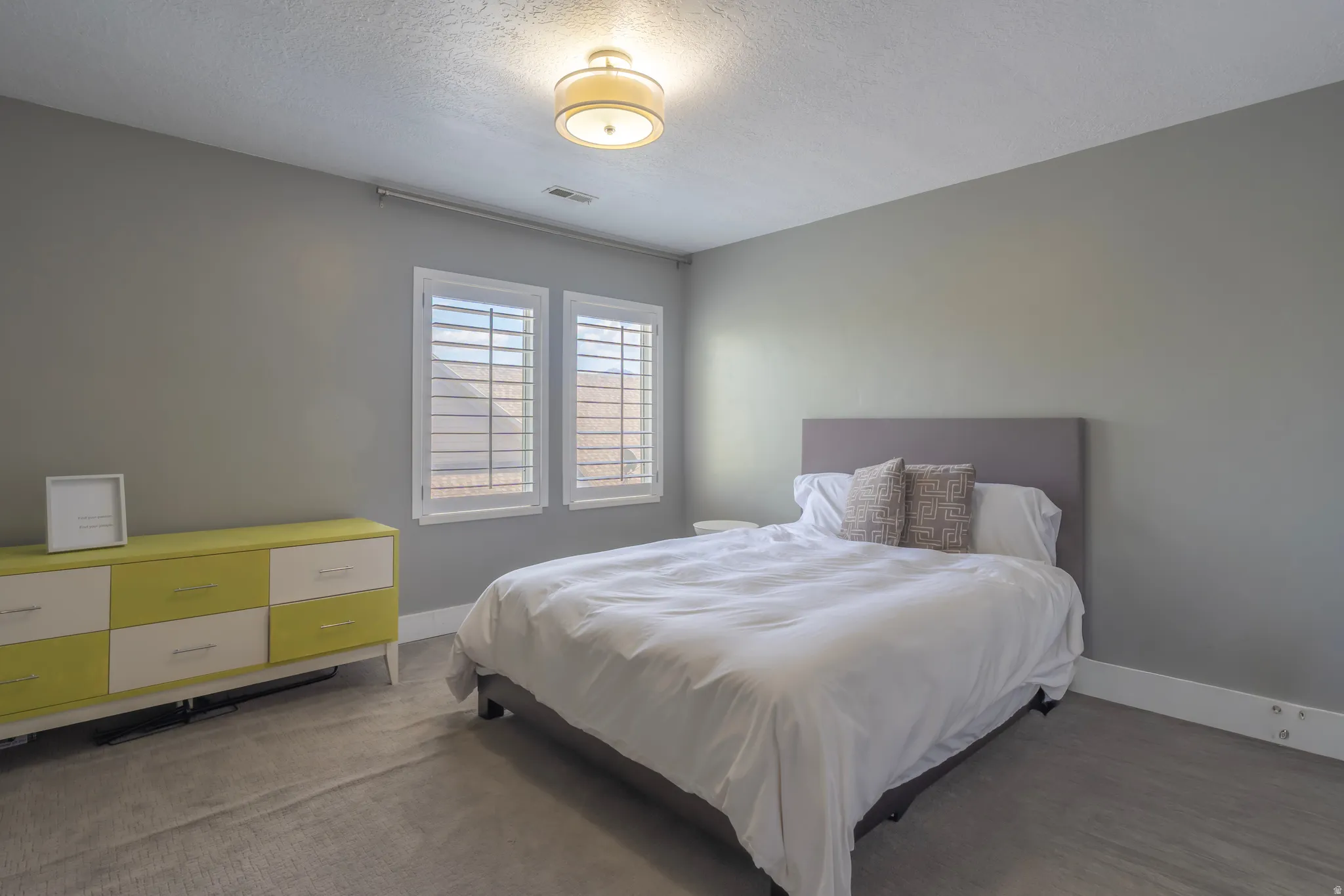 Textured gray wall paint, two windows with plantation shutters, overhead flush-mount light fixture, and light-toned carpet