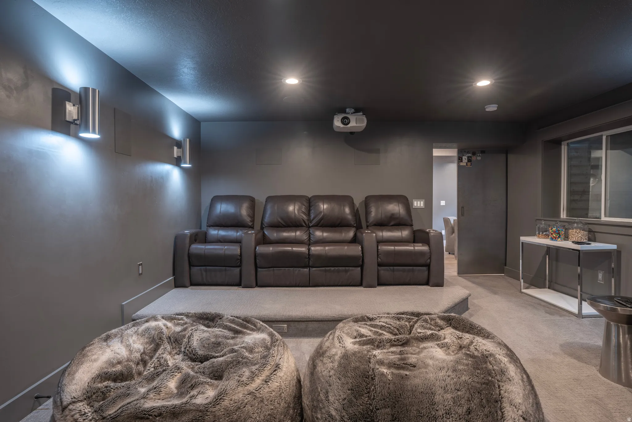 Carpeted media room with a raised platform, wall-mounted sconces, recessed lighting, and a mounted projector