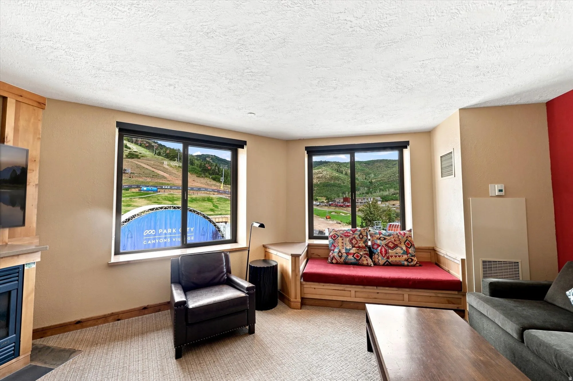 Expansive windows showcasing mountain scenery, built-in wooden daybed, media console with integrated fireplace, and neutral carpeting
