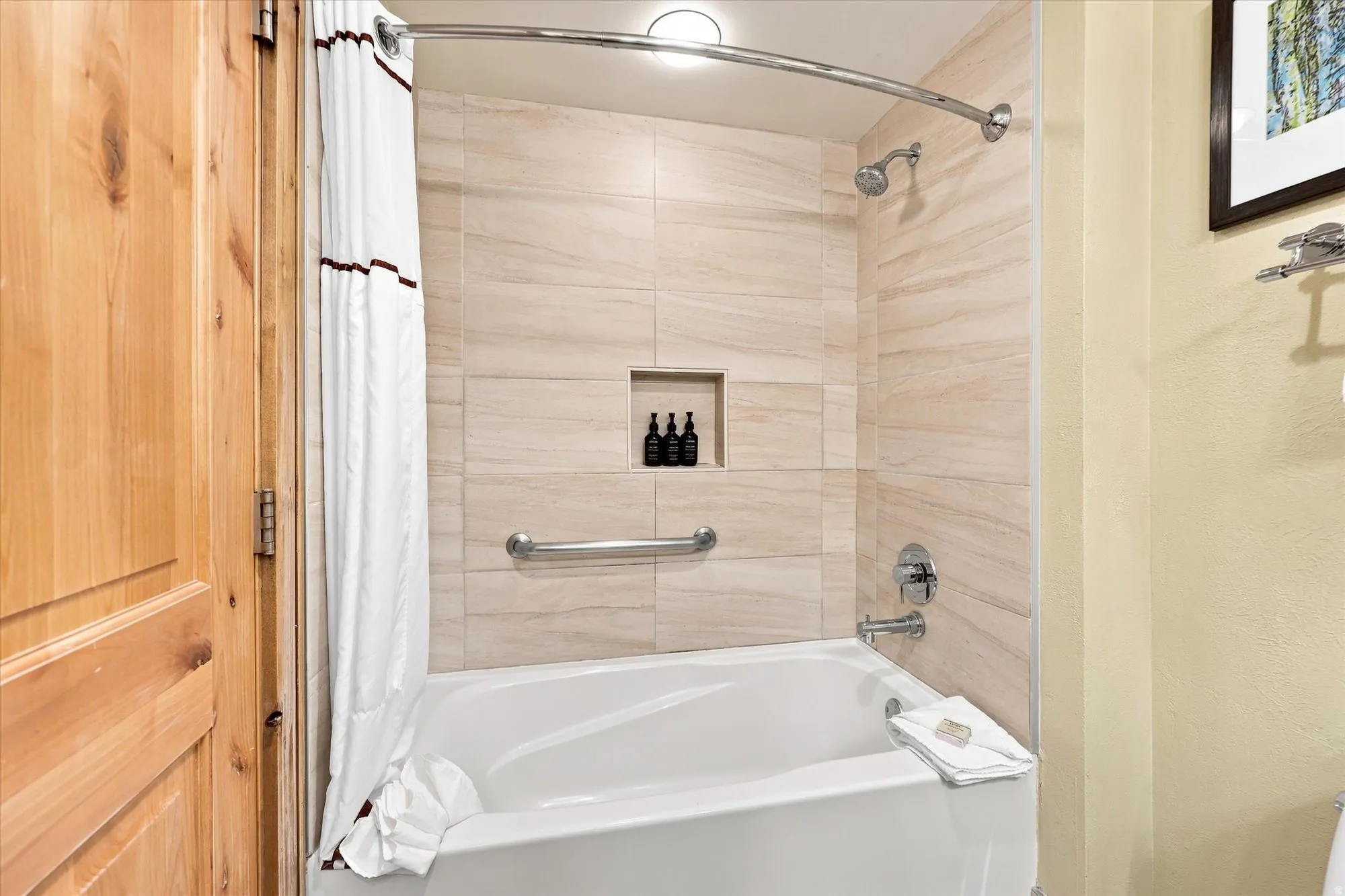 Bathtub with integrated shower, tiled surround, recessed shower niche, chrome fixtures, and a grab bar