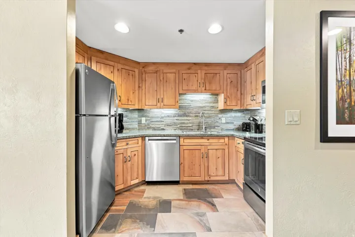 Kitchen featuring wood cabinetry, stainless steel appliances, stone-look tile backsplash, and multi-toned stone tile flooring