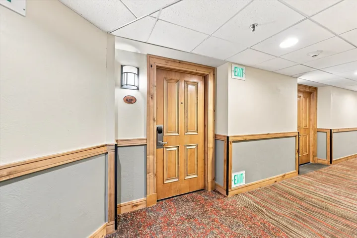 Professional hallway featuring wood-paneled doors with multi-panel detailing, wainscoting with wood-finish rail trim, patterned carpet flooring, and recessed ceiling lighting with emergency exit signage