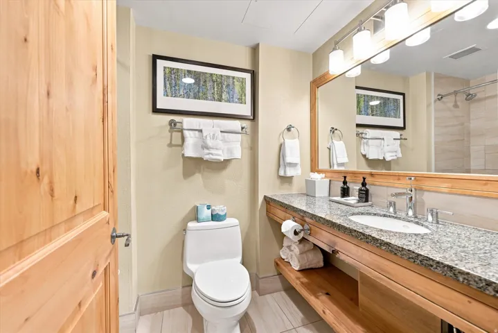 Hardwood door, stone countertop, wood-finish vanity with open shelving, single under-mount sink, and large wall-mounted mirror with integrated lighting
