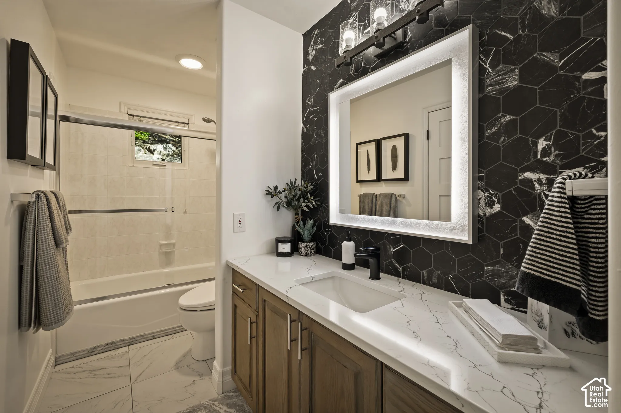 Hexagon tile accent wall, illuminated vanity mirror, wood-finish cabinetry, white countertop with integrated sink, and a black-finish faucet