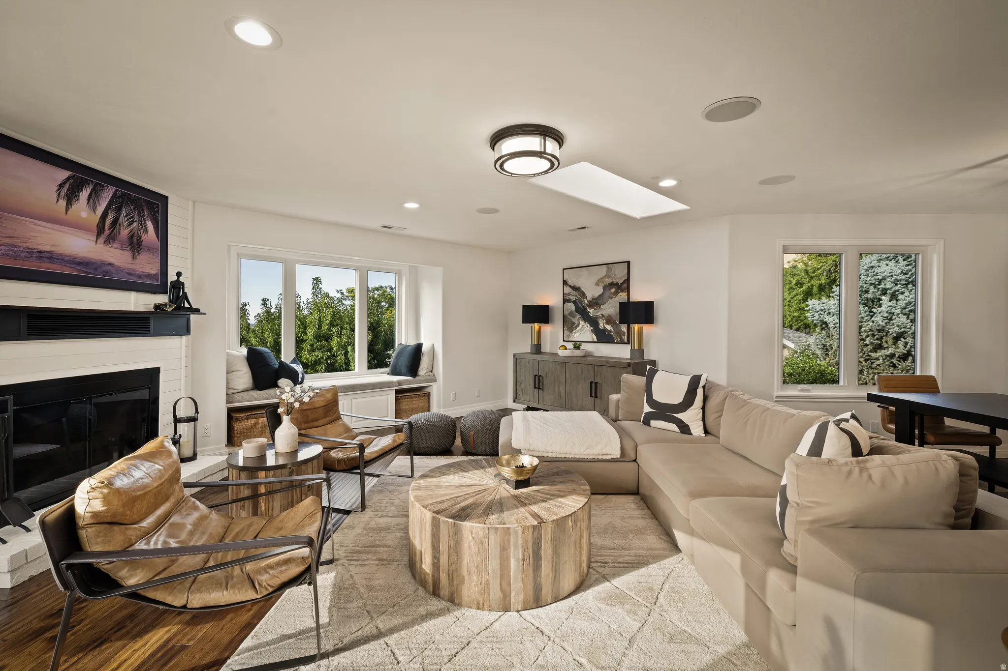White brick fireplace, recessed lighting, expansive bay window with built-in bench seating, wood-finish flooring, and a ceiling-mounted skylight