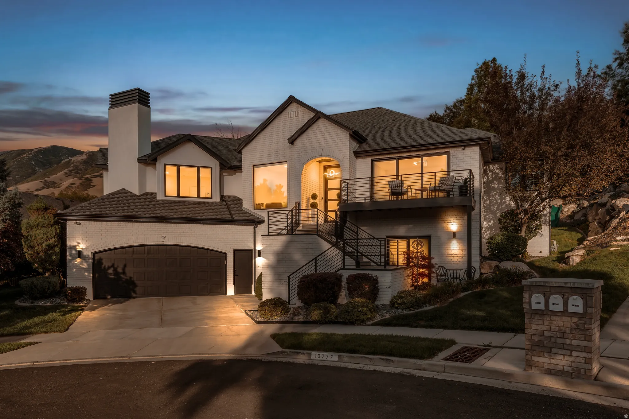 Modern stucco and brick exterior, multi-level design, attached two-car garage, upper-level balcony with metal railings, and a prominent brick chimney feature