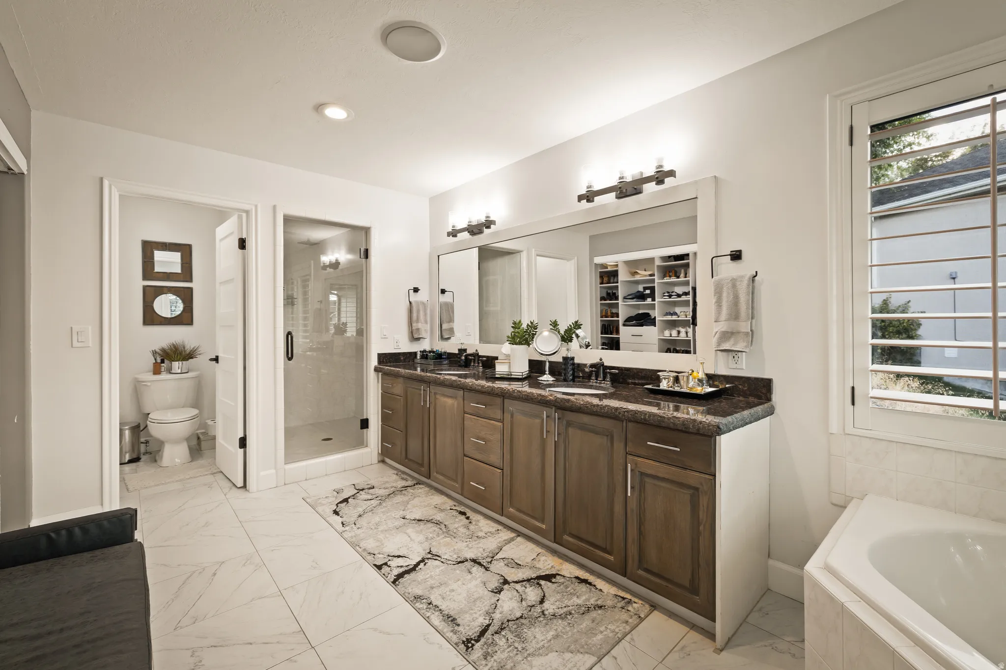 Dual vanity with a dark countertop and wood-finish cabinetry, frameless glass shower enclosure, separate water closet, and a built-in soaking tub