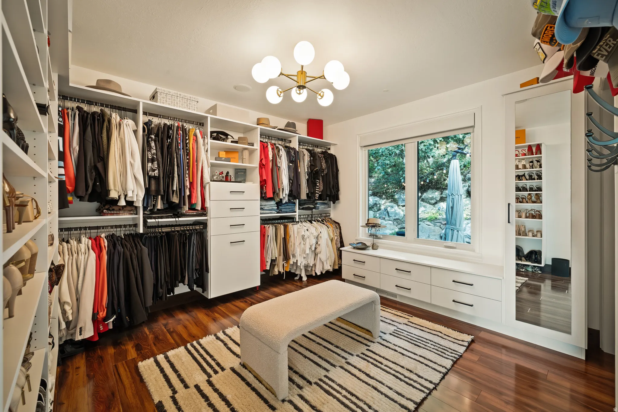 Custom built-in closet systems with integrated drawers, wood-finish flooring, a contemporary ceiling light fixture, a large window, and a full-length mirrored sliding door