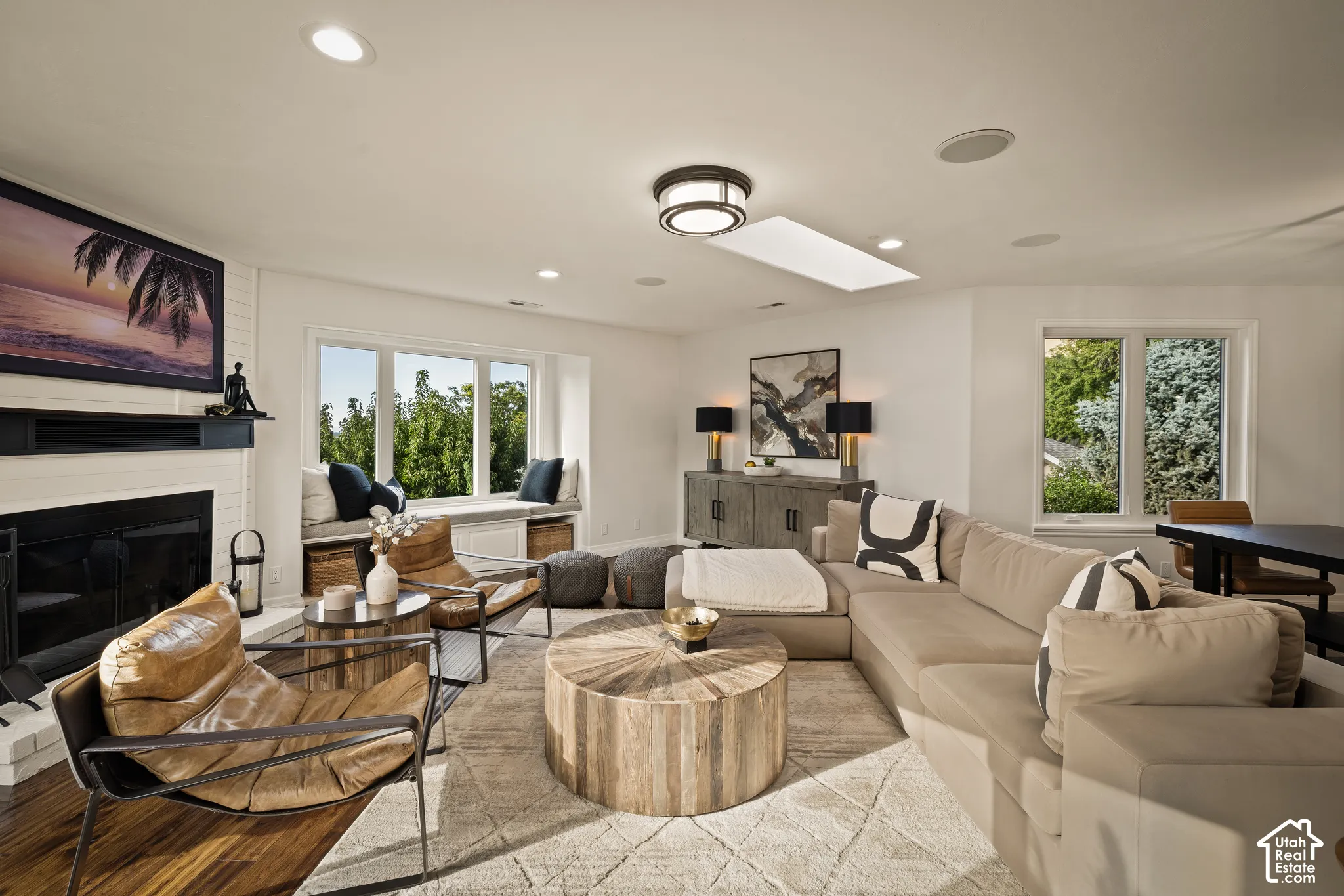 White brick fireplace, recessed lighting, expansive bay window with built-in bench seating, wood-finish flooring, and a ceiling-mounted skylight
