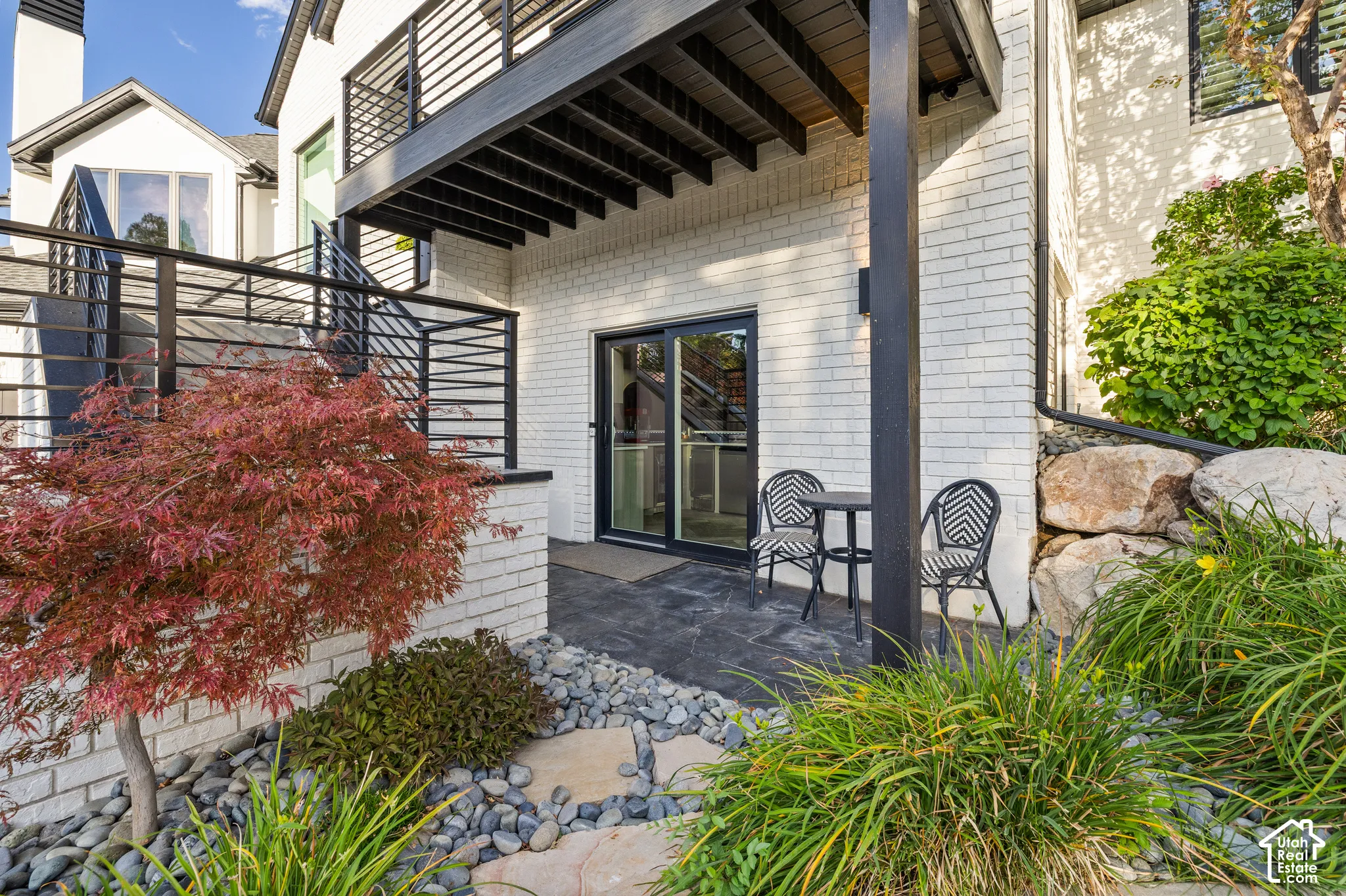 Lower level patio with a sliding glass door, white brick exterior, black horizontal railing, and surrounding landscaping
