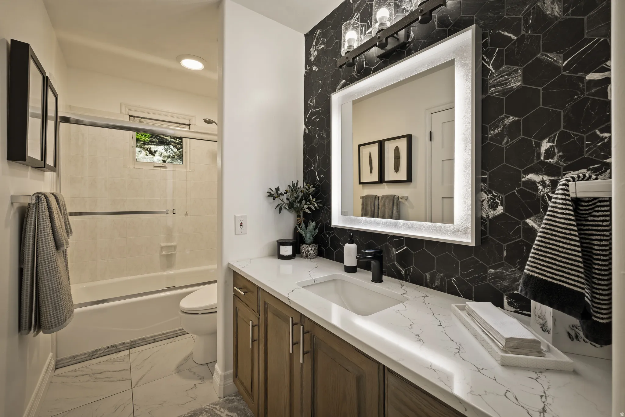 Hexagon tile accent wall, illuminated vanity mirror, wood-finish cabinetry, white countertop with integrated sink, and a black-finish faucet
