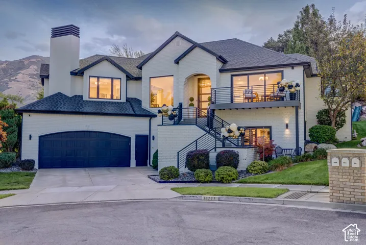 Two-story brick exterior featuring a prominent chimney, an attached multi-car garage, an elevated walk-out balcony with metal railings, and mountain backdrop