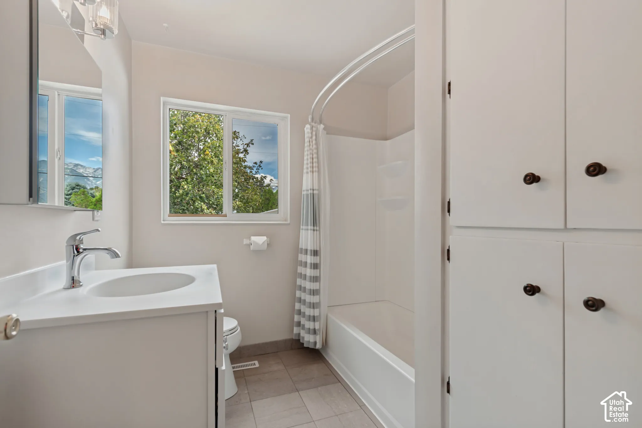 Bathroom featuring a single-sink vanity with white countertop, chrome faucet, window, white built-in cabinetry, and a shower-tub combination. Linen storage and a medicine cabinet mirror