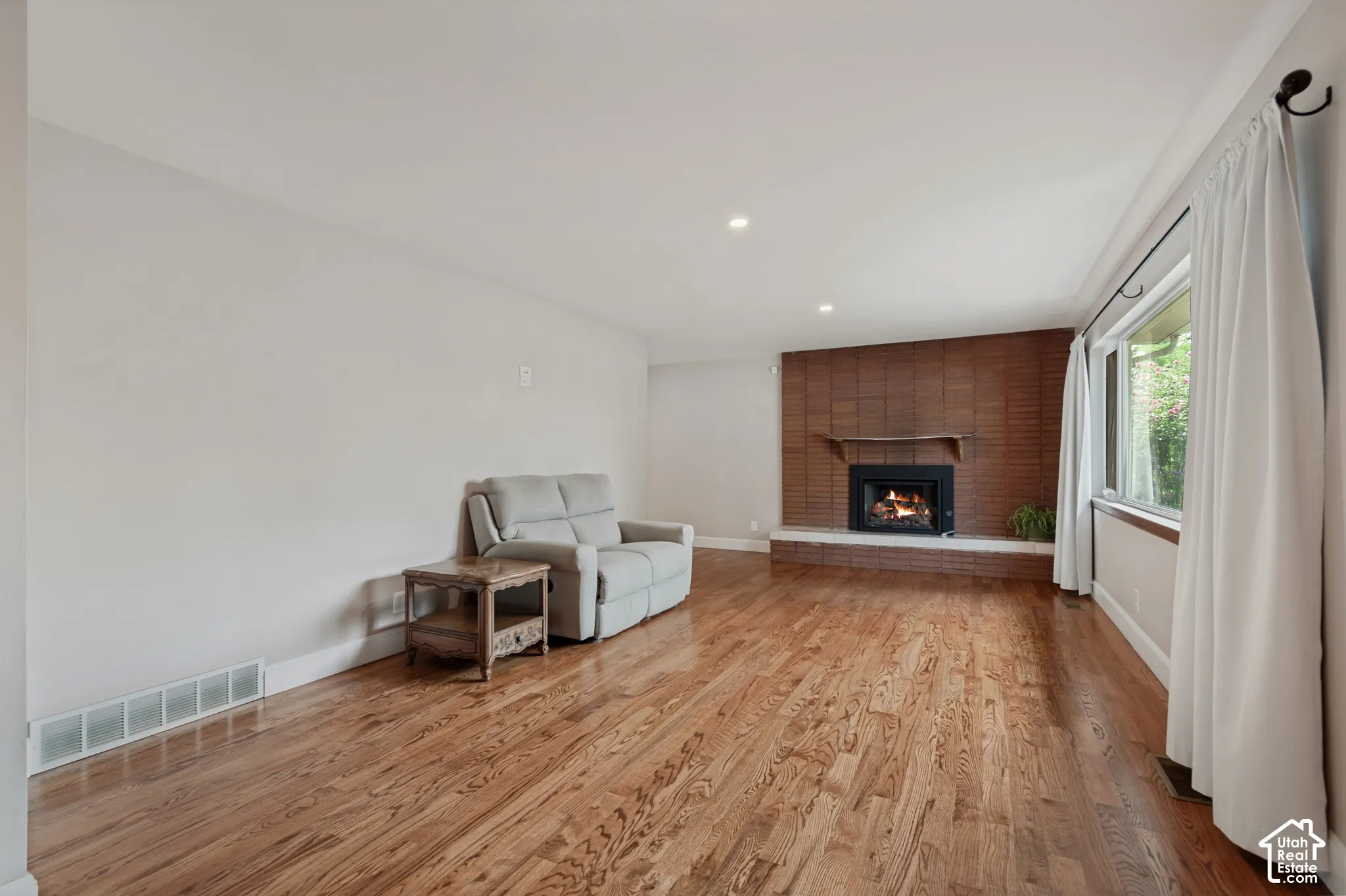 Hardwood flooring, brick gas fireplace with a hearth, large window and recessed lighting