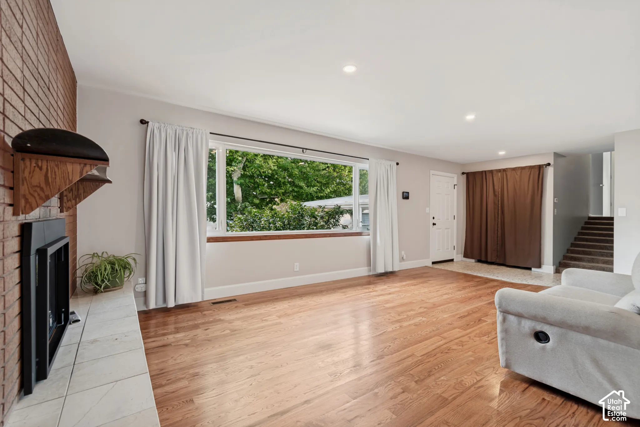 Large picture window, brick-faced fireplace with a tile hearth, wood-finish flooring, and recessed lighting