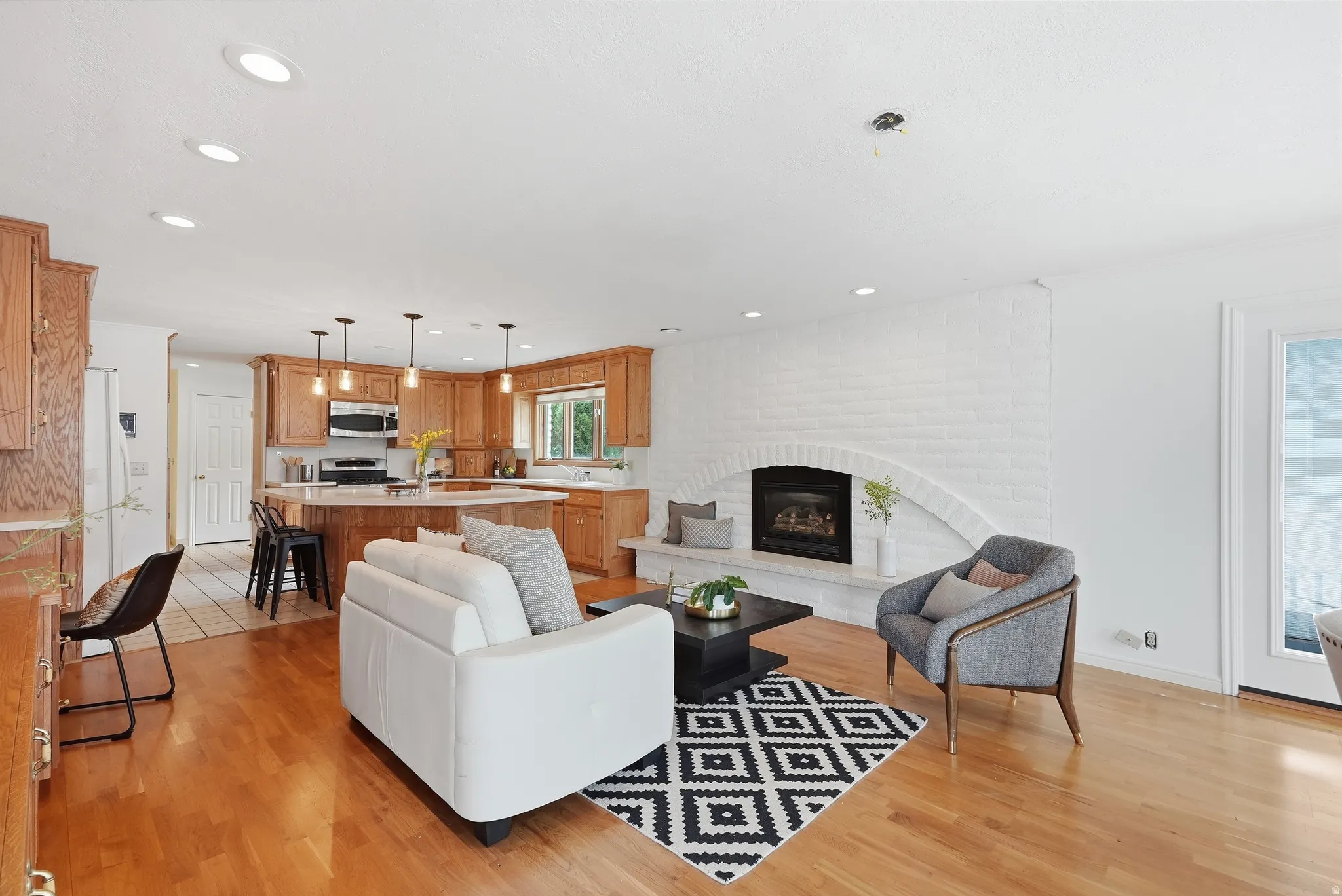Hardwood-style flooring throughout, a white brick fireplace with a built-in hearth, a kitchen island with pendant lighting, recessed lighting, and wood cabinetry