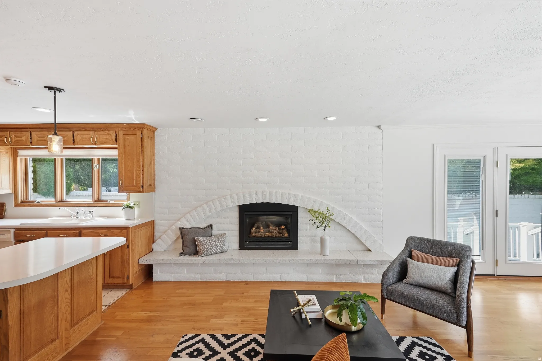 White-painted brick fireplace with an arched hearth, wood-finish flooring, wood cabinetry, built-in kitchen island with white countertop, and glass-paned doors