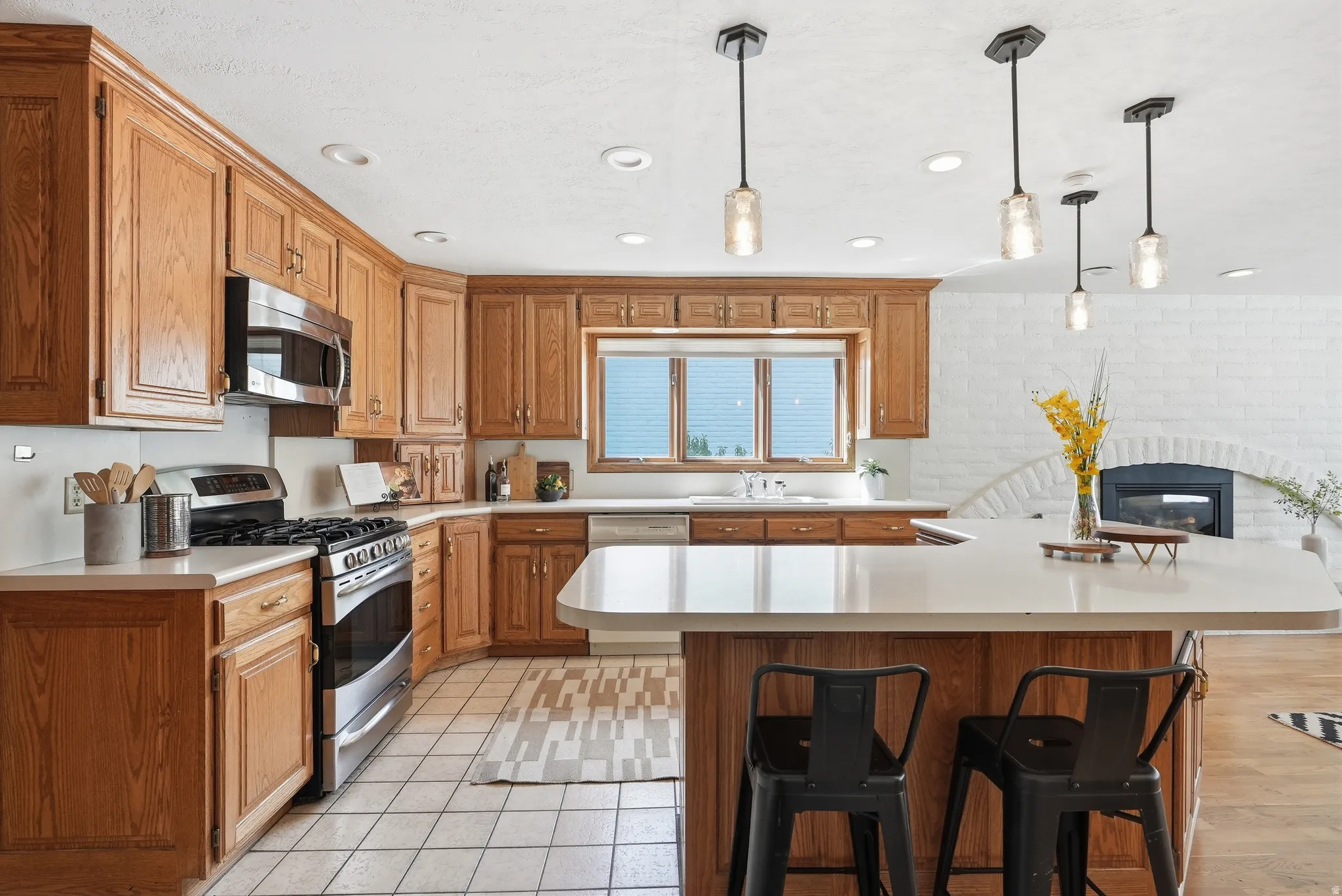Wood-finish cabinetry, stainless steel appliances, a kitchen island with seating, a white brick fireplace, and pendant lighting