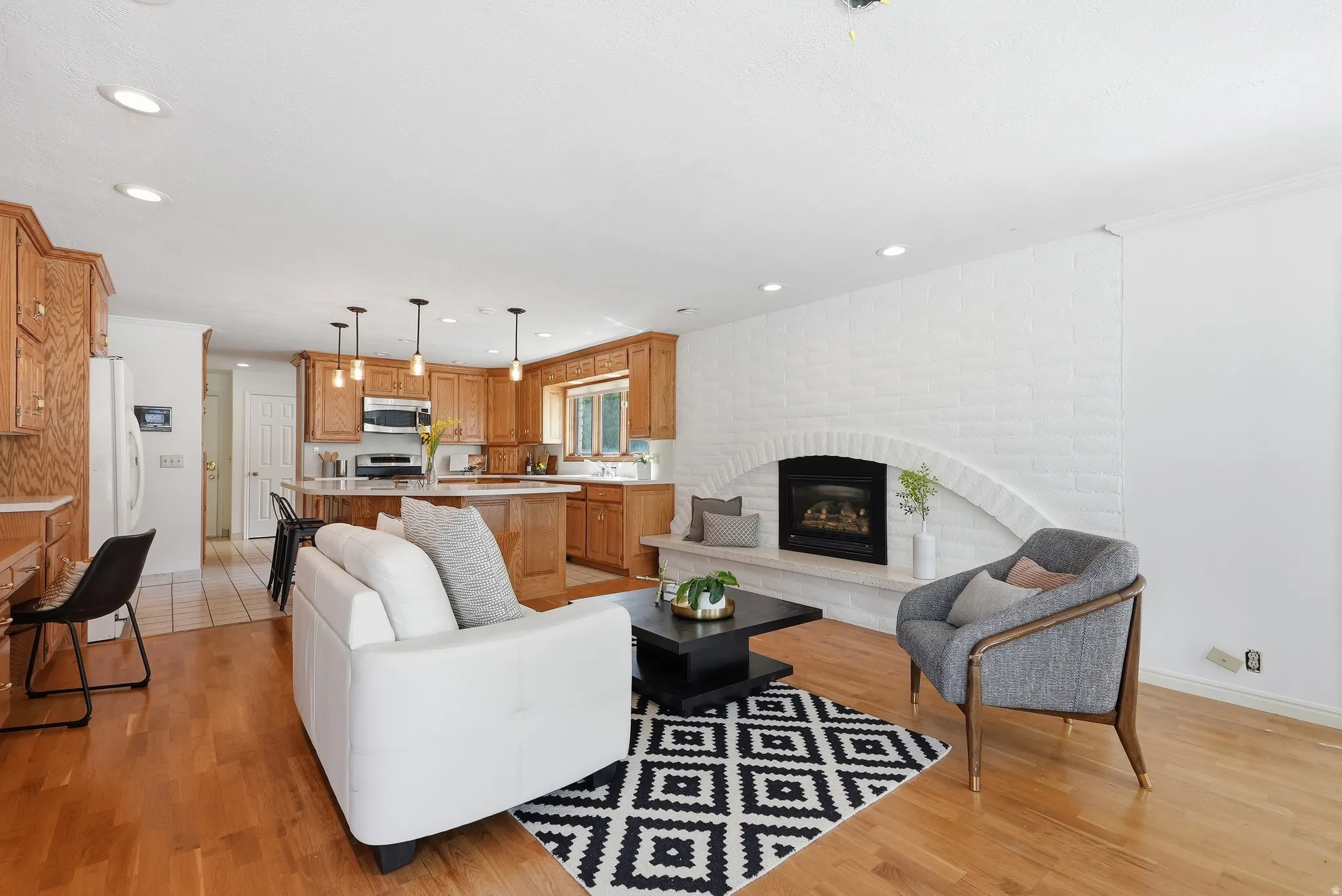 Open-concept living area with wood-finish flooring, a white brick fireplace with an arched hearth, a kitchen island with pendant lighting, and abundant wood cabinetry