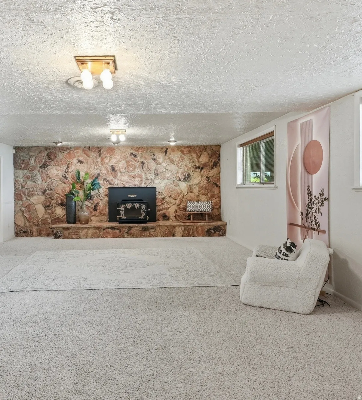 Stone accent wall with wood-burning stove, wall-mounted light fixtures, multiple windows, and light-toned carpeting throughout