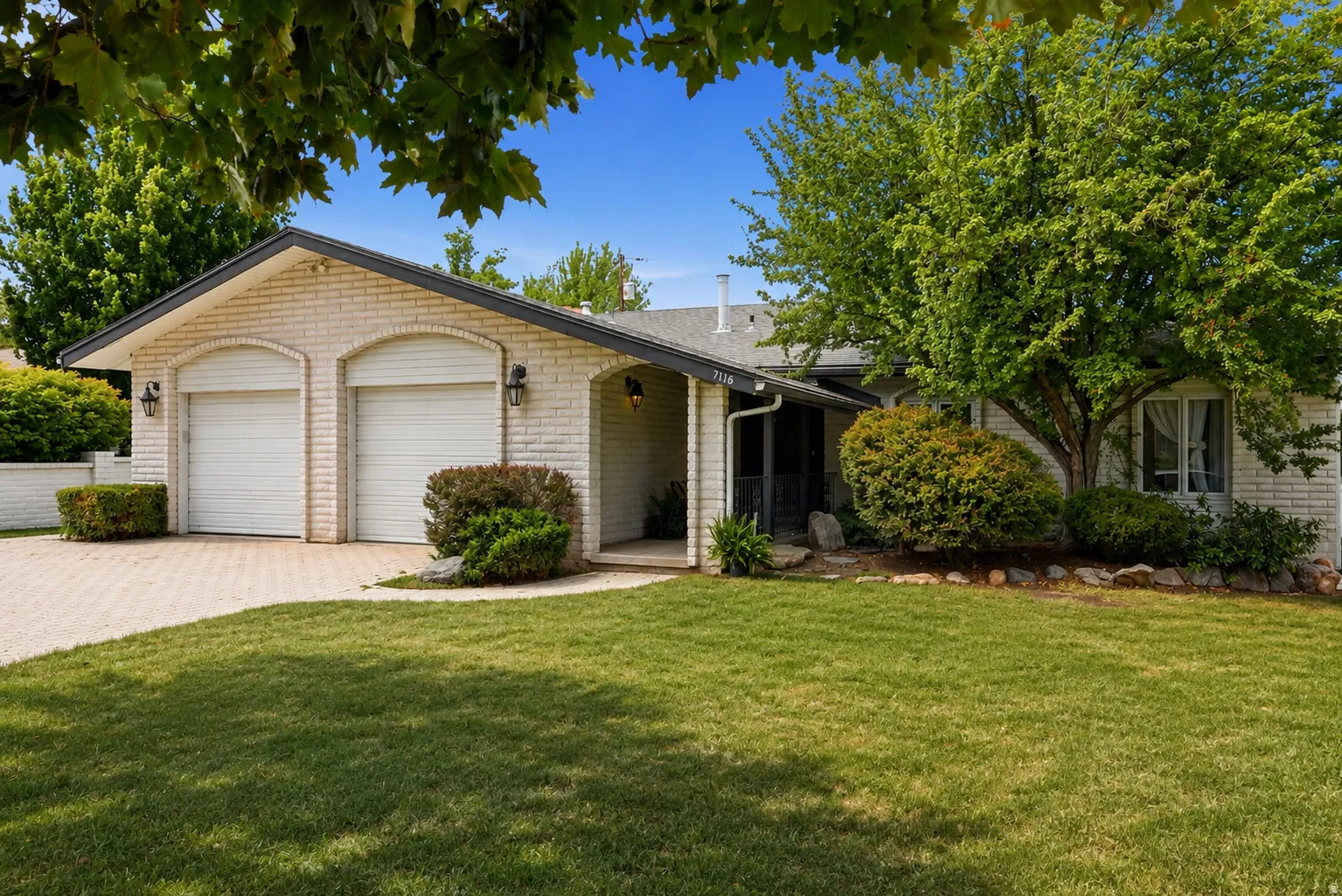 Brick exterior with dual garage doors, paved driveway, mature trees, and established shrubs