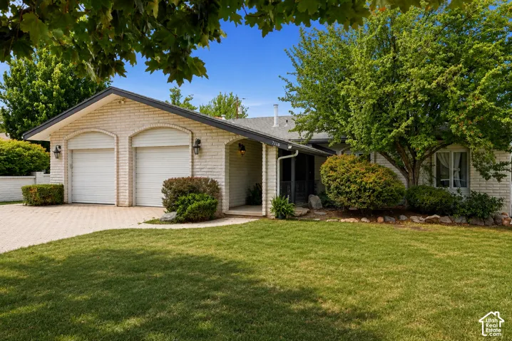 Brick exterior with dual garage doors, paved driveway, mature trees, and established shrubs