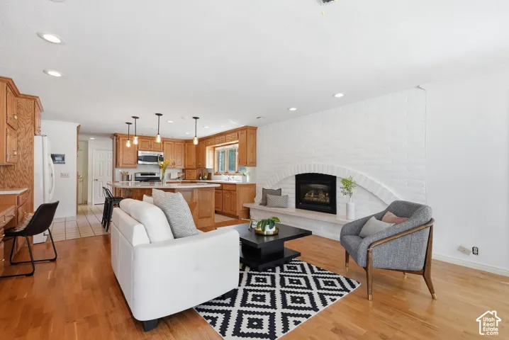 Open-concept living area with wood-finish flooring, a white brick fireplace with an arched hearth, a kitchen island with pendant lighting, and abundant wood cabinetry