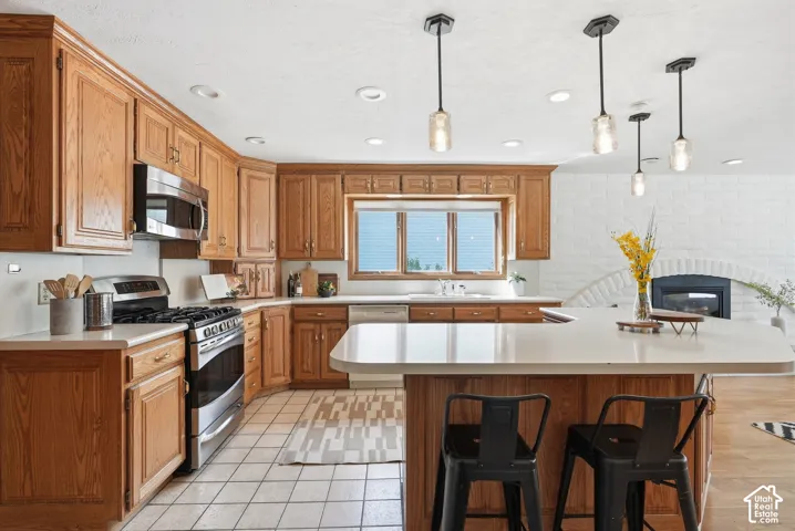 Wood-finish cabinetry, stainless steel appliances, a kitchen island with seating, a white brick fireplace, and pendant lighting
