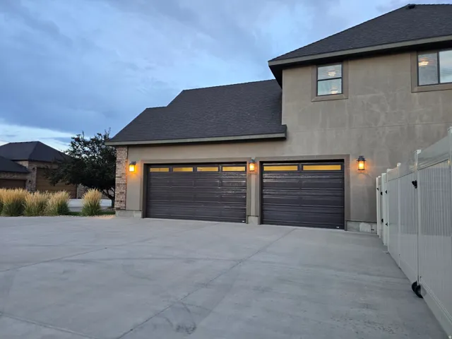 Three-car garage with dark paneled doors and upper window panels, brick veneer accents, exterior sconce lighting, and a concrete driveway