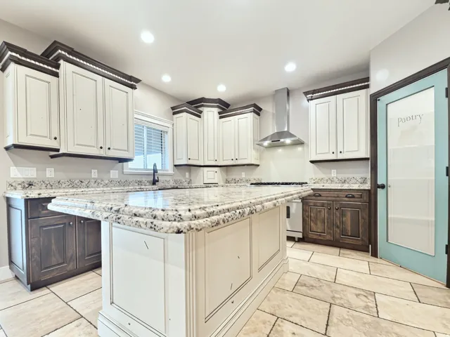Kitchen featuring a central island, granite countertops, two-tone cabinetry, stainless steel range hood, and a pantry door with glass insert