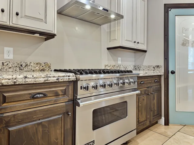 Stainless steel Viking gas range with a built-in oven, granite countertops, wood-finish lower cabinetry, painted upper cabinetry, and tile flooring
