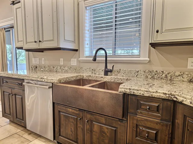 Two-tone cabinetry with light upper and dark lower units, granite countertops, double basin copper farmhouse sink with dark bronze faucet, and stainless steel dishwasher