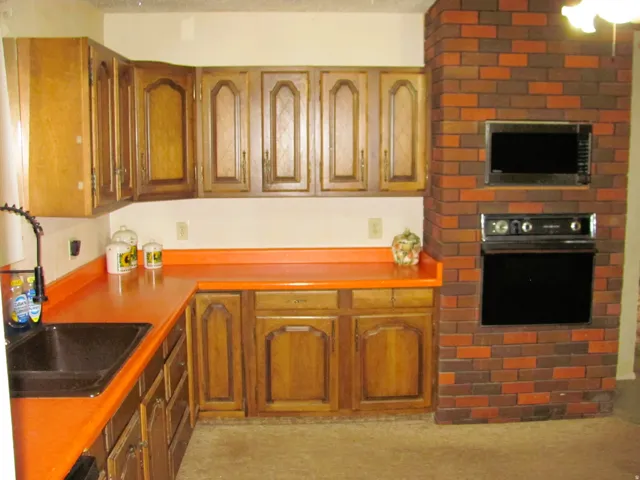Wood-finish cabinetry with routed panel doors, solid surface countertops, built-in microwave, built-in oven, and a brick-finish wall
