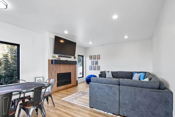 Recessed lighting illuminates wood-finish flooring, a contemporary fireplace with a wood-slat surround, and large black-framed windows