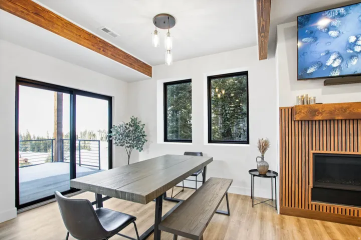 Spacious room featuring exposed wood beams, a built-in fireplace with a slatted wood surround, light wood-finish flooring, and multiple windows with black trim