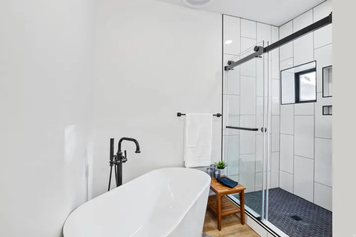 Freestanding soaking tub with a floor-mounted matte black faucet, glass-enclosed shower with black framed sliding door, large format white tiled walls, and a black hex-tiled shower floor