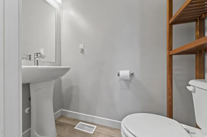 Pedestal sink with chrome faucet, wall-mounted mirror with integrated lighting, wood-finish flooring, and natural wood shelving unit
