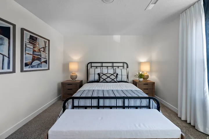 Neutral wall color, textured carpet flooring, ceiling-mounted light fixture, white window treatments, and a black metal bed frame