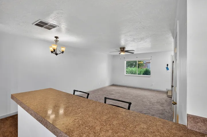 Breakfast bar with a speckled countertop, three-light chandelier, ceiling fan, window with exterior foliage, and wall-to-wall carpeting