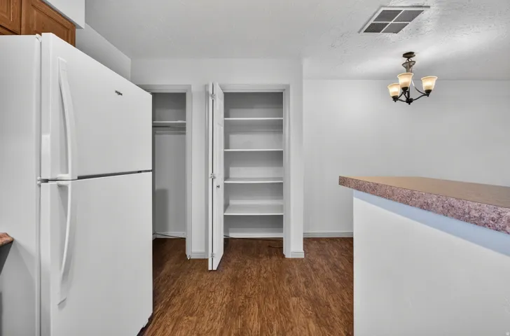 White refrigerator, pantry with shelving, wood-finish flooring, breakfast bar with a speckled countertop, and a four-light chandelier