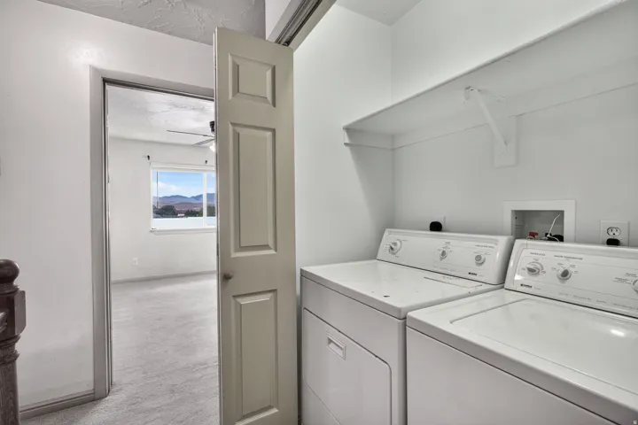 In-unit laundry appliances with overhead shelving, positioned adjacent to a room with carpeting, a ceiling fan, and a window with a mountain backdrop