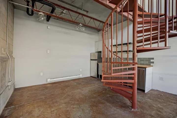 Exposed industrial ceiling, spiral staircase, concrete flooring, kitchen cabinetry, and subway tile backsplash