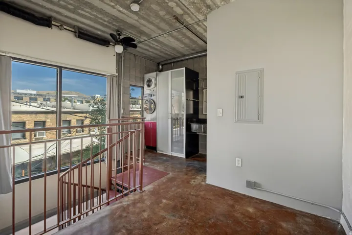 Exposed concrete ceiling, large window with ample natural light, industrial-style staircase, stacked washer and dryer unit, and a built-in kitchen cabinet with a sink