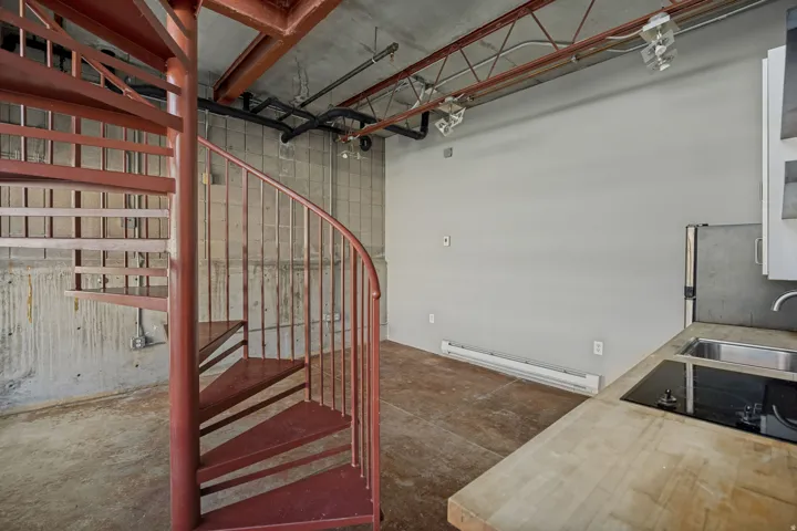 Red metal spiral staircase, concrete flooring, exposed ceiling beams, built-in cooktop, and stainless steel sink